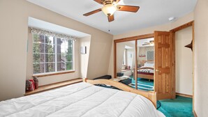 A bedroom with a bed, a ceiling fan, a window with lace curtains, and a wooden door leading to another room.