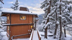 A wooden cabin with a staircase leading to a deck, surrounded by snow-covered trees.