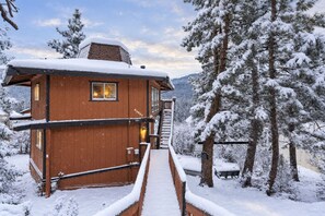A wooden cabin with a staircase leading to a deck, surrounded by snow-covered trees.
