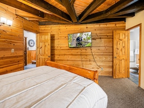 Quarto de cabana de madeira com cama, televisão fixada na parede e porta que dá para outro cômodo.