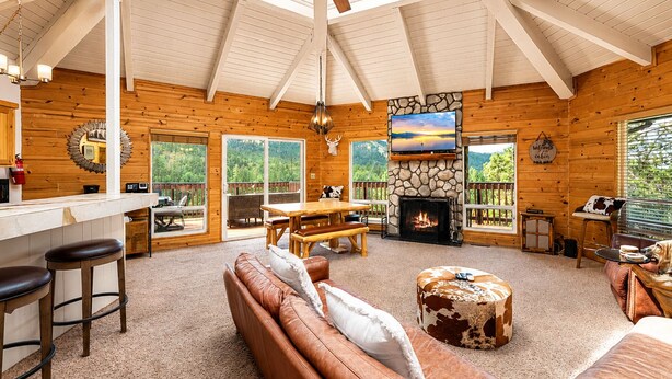 A cabin interior with a fireplace, wooden furniture, a stone accent wall, and a large window overlooking greenery.