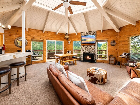 A cabin interior with a fireplace, wooden furniture, a stone accent wall, and a large window overlooking greenery.
