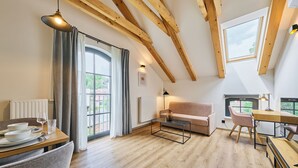 Een ruime woonruimte met een houten plafond, een bank, een eettafel met stoelen en een groot raam dat voor natuurlijk licht zorgt.