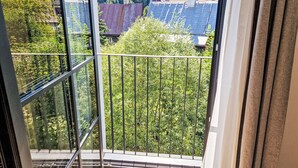 Een balkon met een metalen hekwerk, een glazen deur en uitzicht op een groen landschap met bomen en gebouwen.