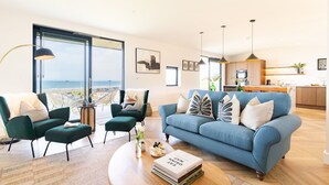 A modern living room with a view of the beach, a blue sofa, green chairs, and a dining area with a kitchen in the background.
