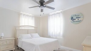 Une chambre à coucher comprenant un lit, une table de chevet, une commode, un ventilateur de plafond et une fenêtre avec des rideaux.