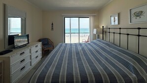 A bedroom with a bed, a dresser, a chair, a TV, and a view of the beach through a large window. - Ocean Cove 108 - Oceanfront in Garden City Beach (Garden City)