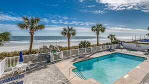 A pool area with a view of the beach, palm trees, and a fenced deck with chairs and umbrellas. - Ocean Cove 108 - Oceanfront in Garden City Beach (Garden City)