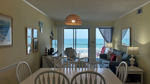 A dining area with a table and chairs, a sofa, a lamp, and a view of the ocean through large windows. - Ocean Cove 108 - Oceanfront in Garden City Beach (Garden City)