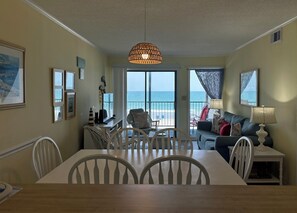 A dining area with a table and chairs, a sofa, a lamp, and a view of the ocean through large windows. - Ocean Cove 108 - Oceanfront in Garden City Beach (Garden City)