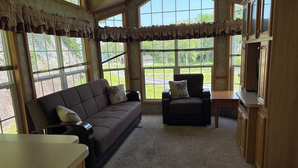 Una sala de estar acogedora con un sofá y un sillón color café, una mesita y un televisor. Amplias ventanas ofrecen vistas al exterior.