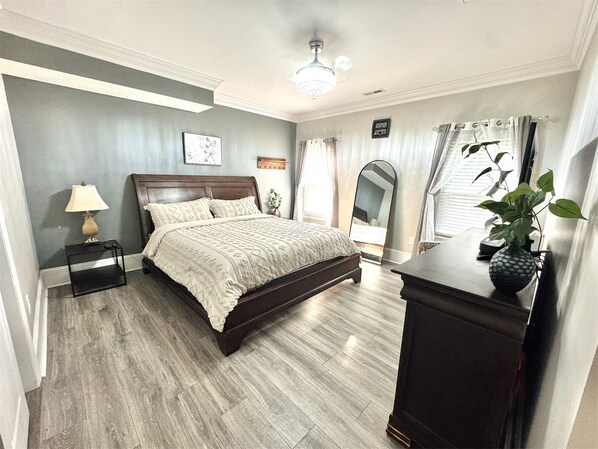 Un dormitorio con cama, mesita de noche, lámpara, espejo, planta en maceta y cómoda.