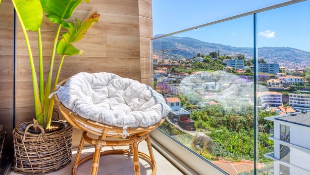 A balcony with a wicker chair and a potted plant, overlooking a cityscape with buildings and hills. - The Place Funchal View, a Home in Madeira (Funchal)