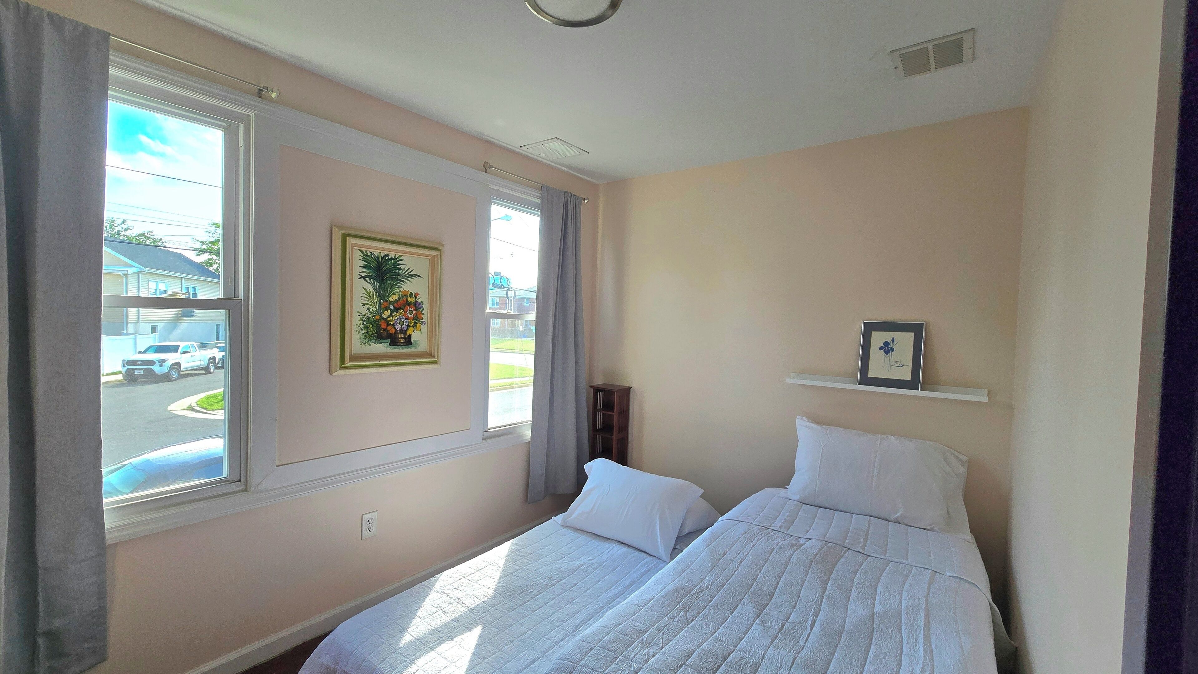 A bedroom with two beds, a window with a view of a street, a framed floral artwork, and a small shelf with a picture.