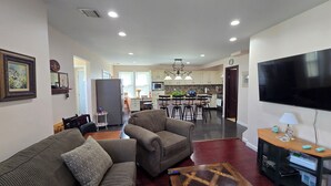 A modern living room with a sofa, a coffee table, and a TV. There is a kitchen in the background with a dining area.