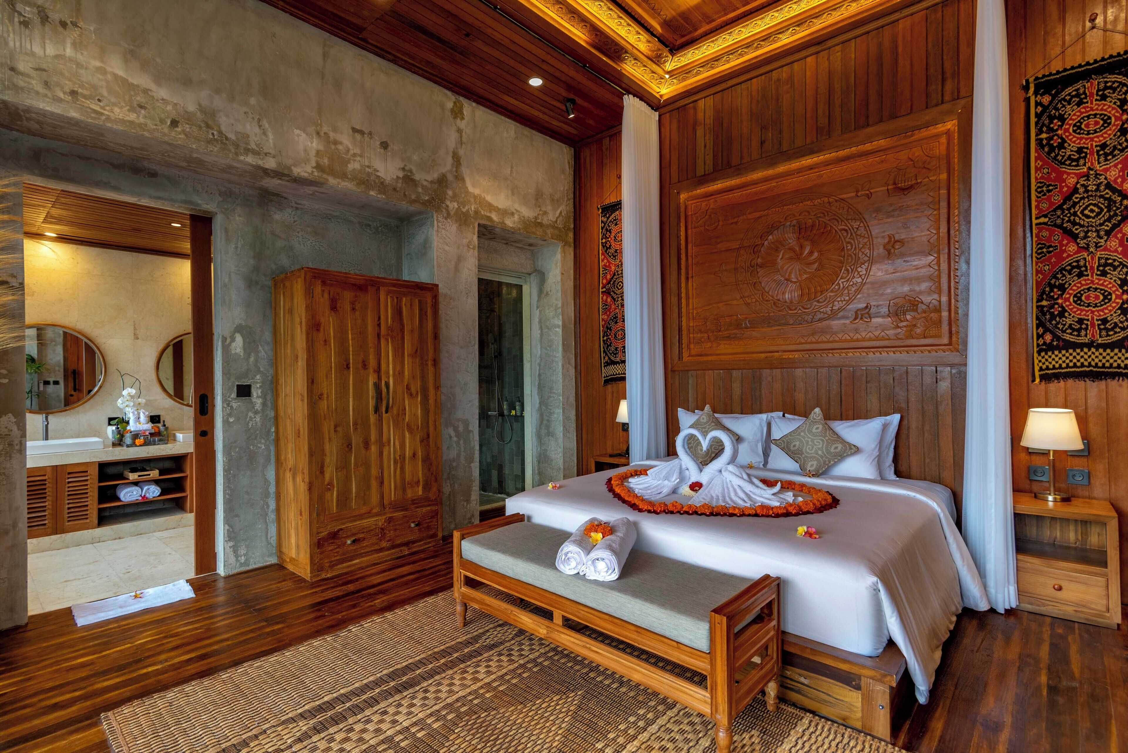 Ein Schlafzimmer mit einem hölzernen Bett, einer Sitzbank, einem Badezimmer mit Waschbecken und einem Spiegel.