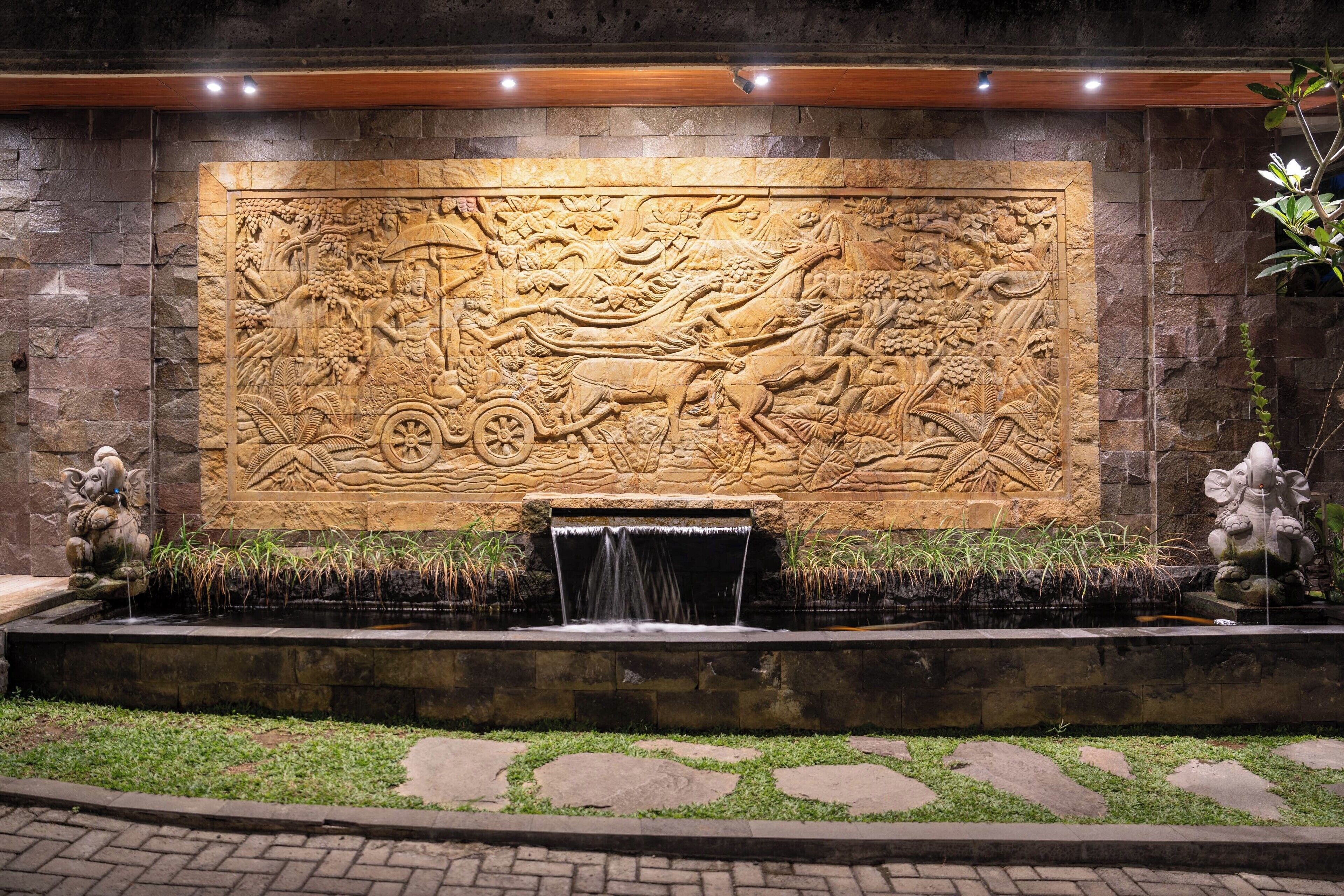 A stone wall with a detailed bas-relief carving, a small water feature, and two statues.