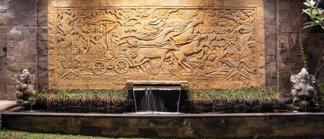 A stone wall with a detailed bas-relief carving, a small water feature, and two statues.