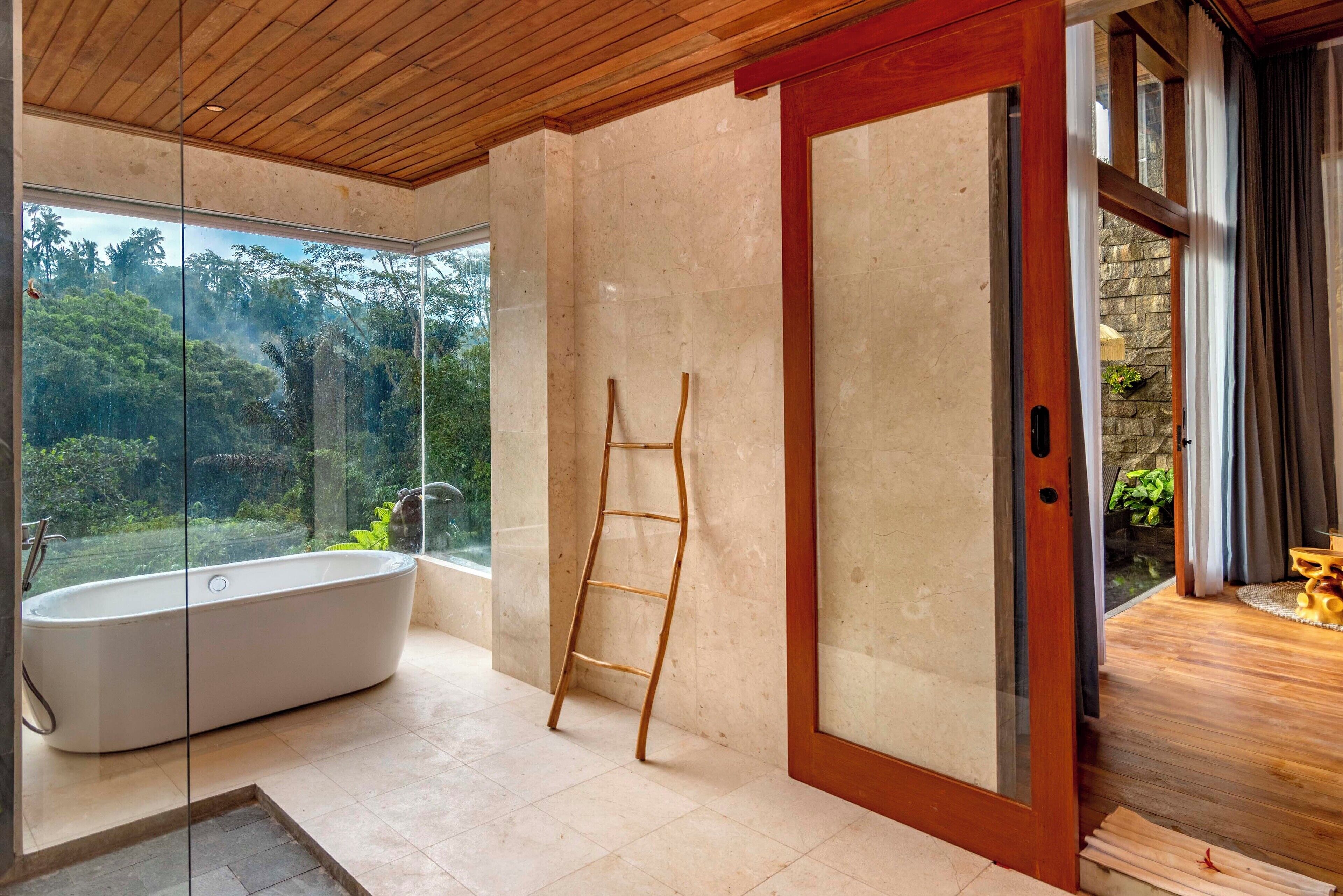 Un baño moderno con una bañera independiente, una escalera de madera y un cristal que da a una pared de piedra.