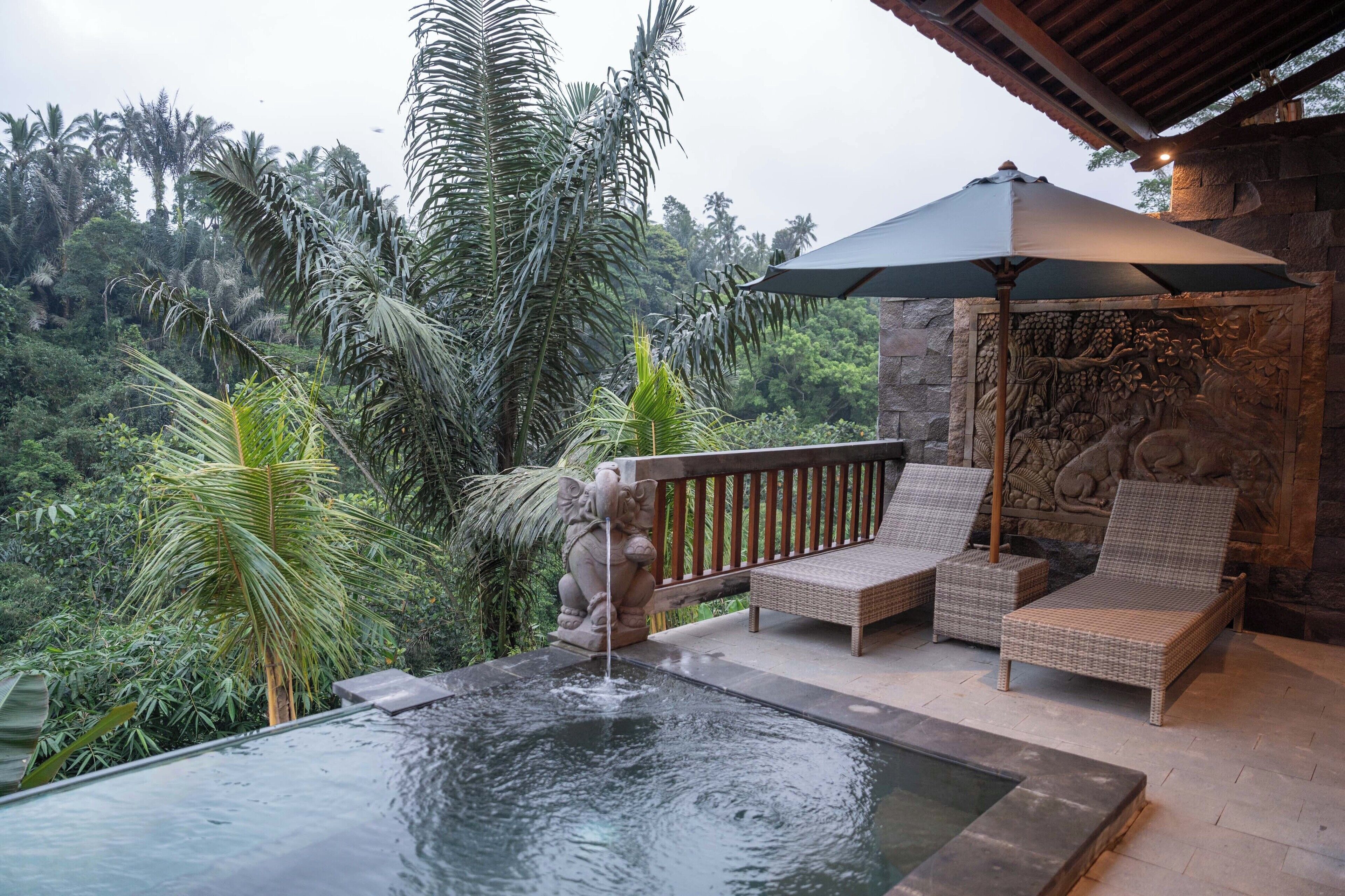 A serene outdoor seating area with a stone wall, a water feature, and lush greenery in the background.