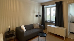 Sala de estar com sofá, uma pequena mesa, luminária, radiador, janela com cortinas e um espaço com cozinha compacta.