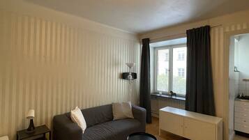 Una sala de estar con sofá, una mesita, lámpara, radiador, ventana con cortinas y una zona de kitchenette.