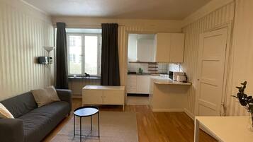 Un espacio habitable pequeño con un sofá, una mesita redonda, una kitchenette y una puerta.