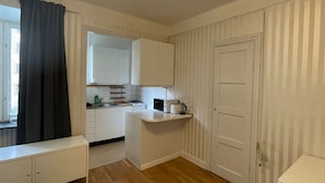 Cozinha compacta com armários brancos, bancada e uma pequena ilha. Há uma janela com cortina preta, uma porta e um banco branco em primeiro plano.