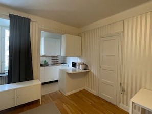 Eine kleine Küche mit weißen Schränken, einer Arbeitsplatte und einer kleinen Kochinsel. Ein Fenster mit schwarzem Vorhang, eine Tür und eine weiße Bank befinden sich im Vordergrund.