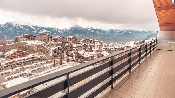 A view from a balcony overlooking a town with snow-covered buildings and mountains in the background.