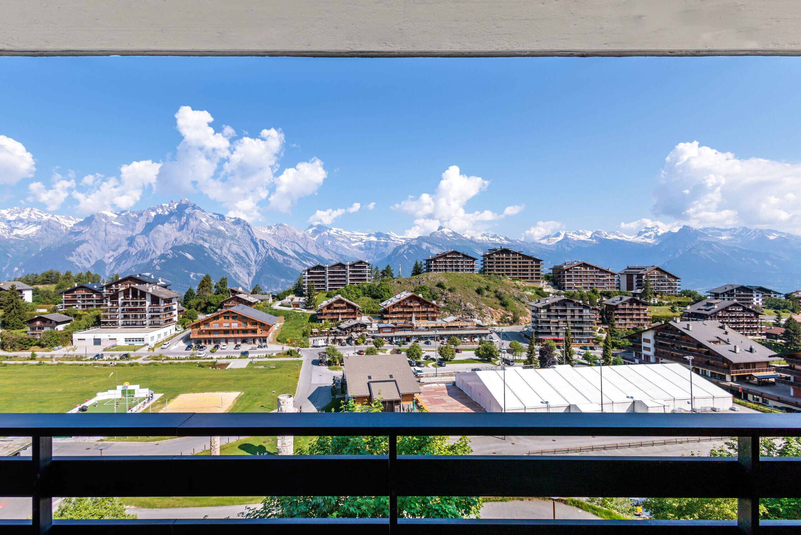 A mountain resort with multiple chalet-style buildings, a ski slope, and snow-capped mountains in the background.