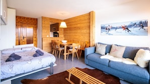 A cozy room with a bed, a blue sofa, a wooden dining table with chairs, and a painting of skaters on the wall. - Christiania II S2 - 4 pers - près des pistes (Haute-Nendaz)