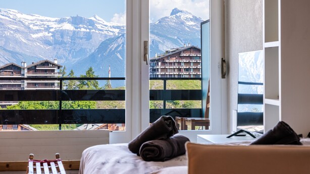 A hotel room with a large window offering a mountain view, a bed with a rolled towel, and a small wooden stool. - Christiania II S2 - 4 pers - près des pistes (Haute-Nendaz)