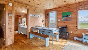 A wooden cabin interior with a dining table, bench, and a bed in the background.