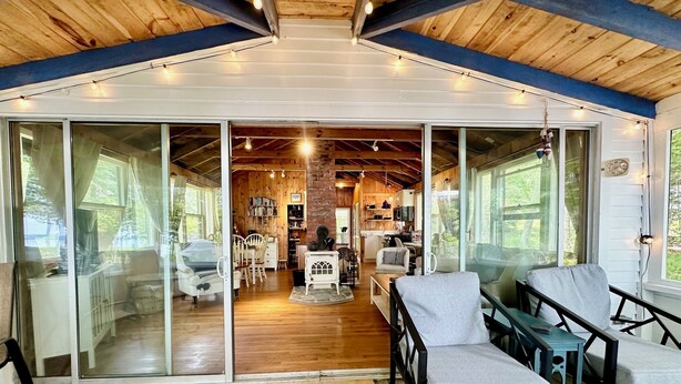 A spacious interior with a wooden floor, a central seating area with chairs and a sofa, a kitchen area, and large sliding glass doors leading to an outdoor deck. - Seaside Cabin Retreat – Breathtaking Views & Ocean Access (Harpswell)