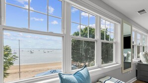 Una habitación con grandes ventanales con vistas a la playa, una cama con un almohadón azul y un aparador.