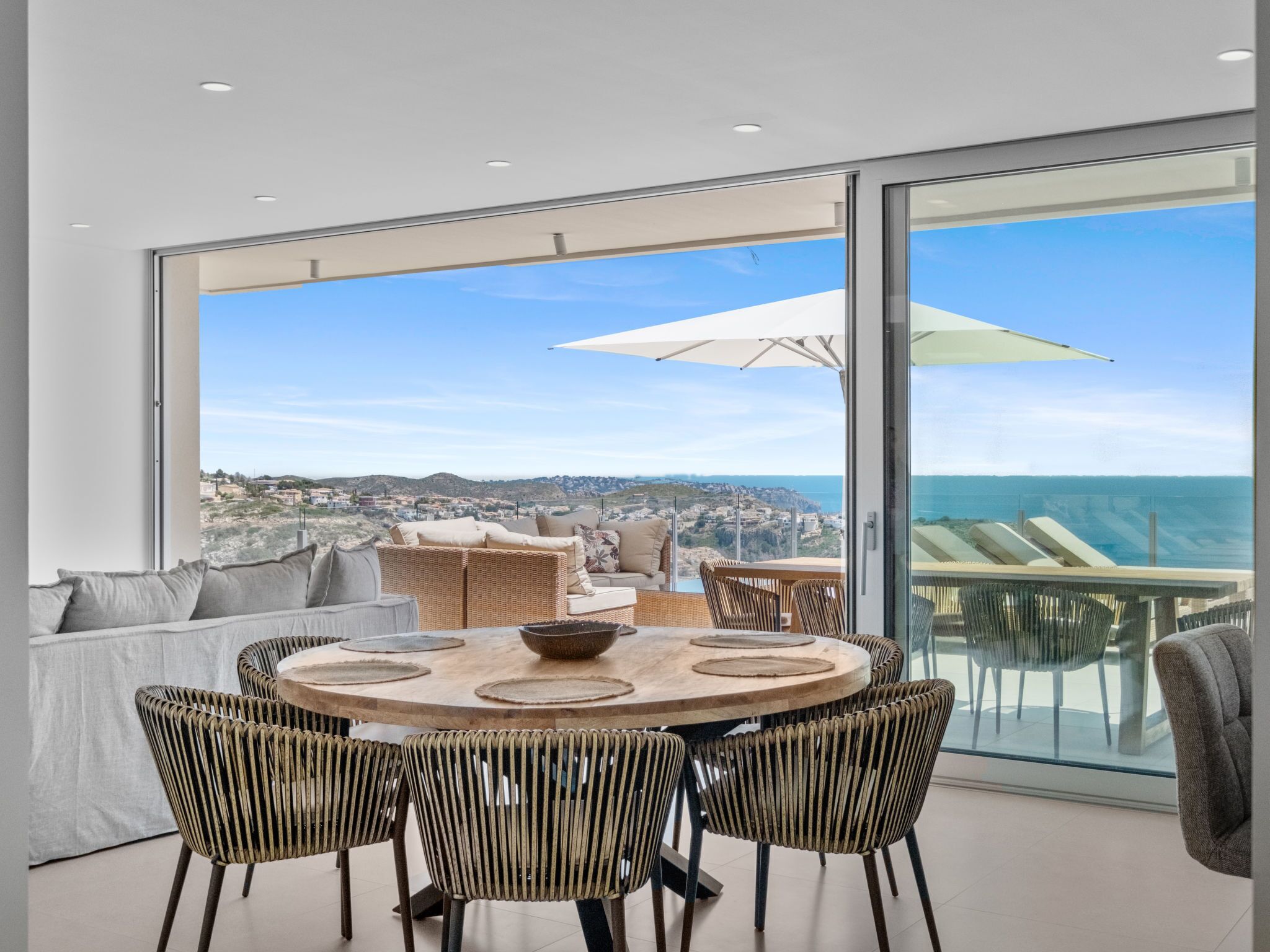 Uma área de refeições moderna com uma mesa redonda, cadeiras de vime e vista para o oceano e para a cidade através de amplas janelas.