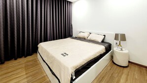 A modern bedroom with a bed, dark curtains, a bedside lamp, and a wooden floor.