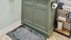 A bathroom with a white tiled wall, a green vanity, a mirror, a towel rack, and a shelf with toiletries. - Chic Casita with AC, WiFi in peaceful Coachella (Coachella)