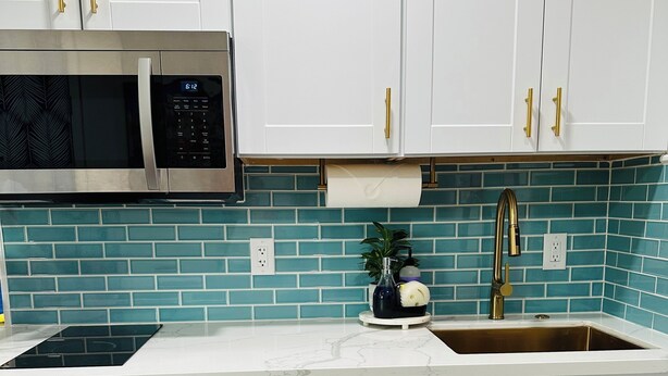 A kitchen with white cabinets, a stainless steel microwave, a sink with a gold faucet, and teal-tiled backsplash. - Chic Casita with AC, WiFi in peaceful Coachella (Coachella)