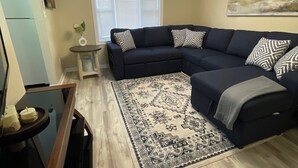 A living room with a dark-colored sectional sofa, a patterned rug, a small side table, and a window with blinds. - The C&C
Comfort and Convenience (Milwaukee)
