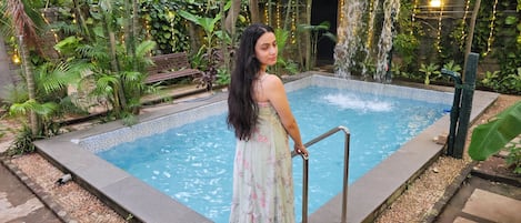 Een vrouw staat bij een zwembad met een waterval, omringd door weelderige begroeiing en een stenen pad.