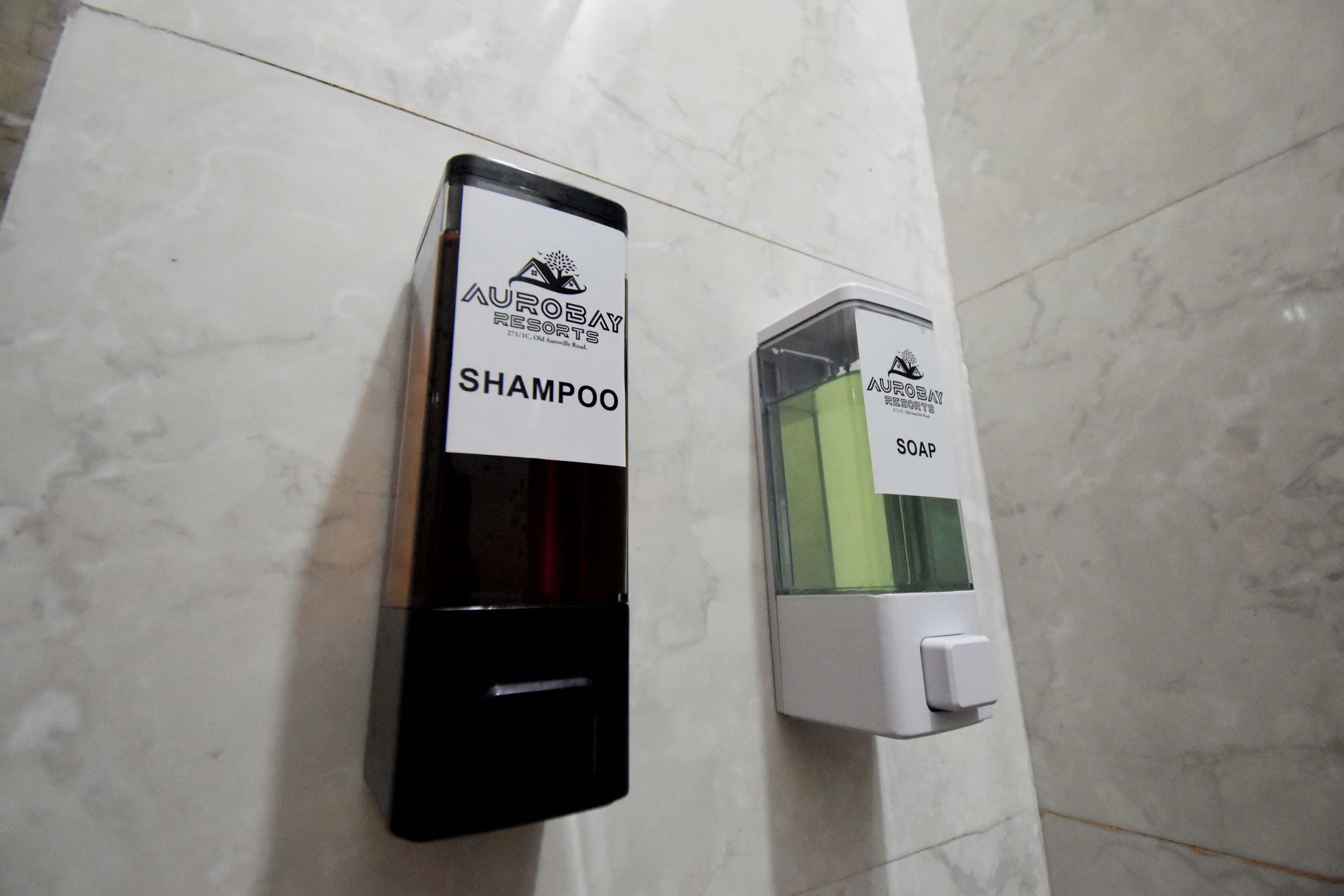 Un dispensador de pared con una botella negra etiquetada 'AuroBay Resorts Shampoo' y un dispensador blanco con una botella verde etiquetada 'AuroBay Resorts Soap'.