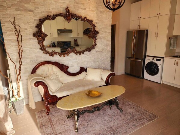 Un salon comprenant un canapé vintage, une table basse, un grand miroir et un coin cuisine avec des placards et des appareils électroménagers.