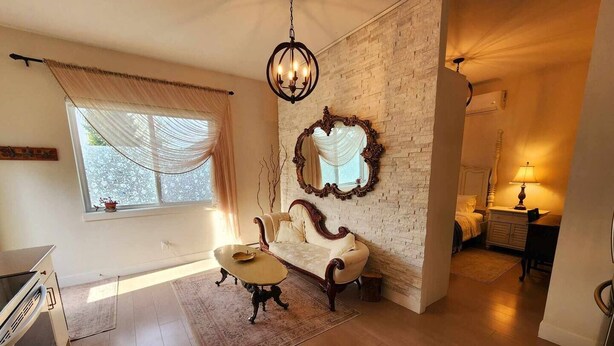 A spacious living room with a white sofa, a wooden coffee table, and a large mirror on a brick wall. There is a window with sheer curtains and a chandelier hanging from the ceiling.