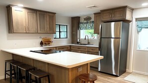 A modern kitchen with wooden cabinets, a stainless steel refrigerator, a large island with stools, and a window with blinds. - Lake Front Cottage | Kayaks | Game Table Fun (Shipshewana)