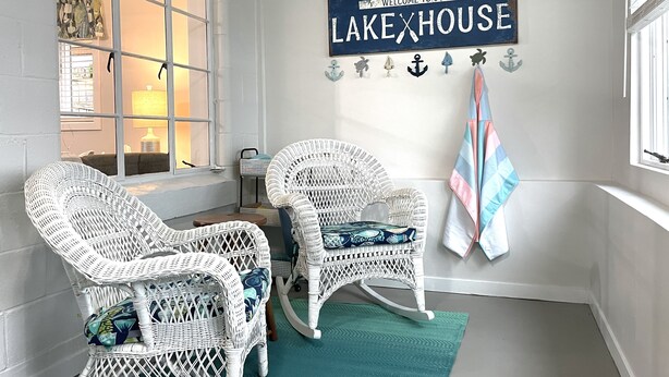 A cozy room with wicker chairs, a rocking chair, a teal rug, and a sign reading "Welcome to our Lake House." - Lake Front Cottage | Kayaks | Game Table Fun (Shipshewana)