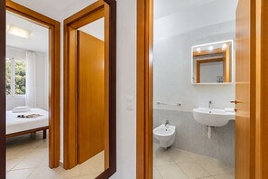 Ein Hotelzimmer mit Bett, Nachttisch, Fenster mit Vorhängen sowie einem Badezimmer mit Waschbecken, Bidet und Spiegel.