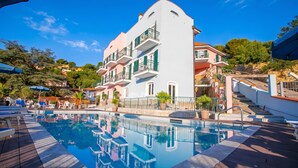 Une piscine avec une terrasse en bois, un bâtiment blanc doté de balcons et un ciel bleu azur.