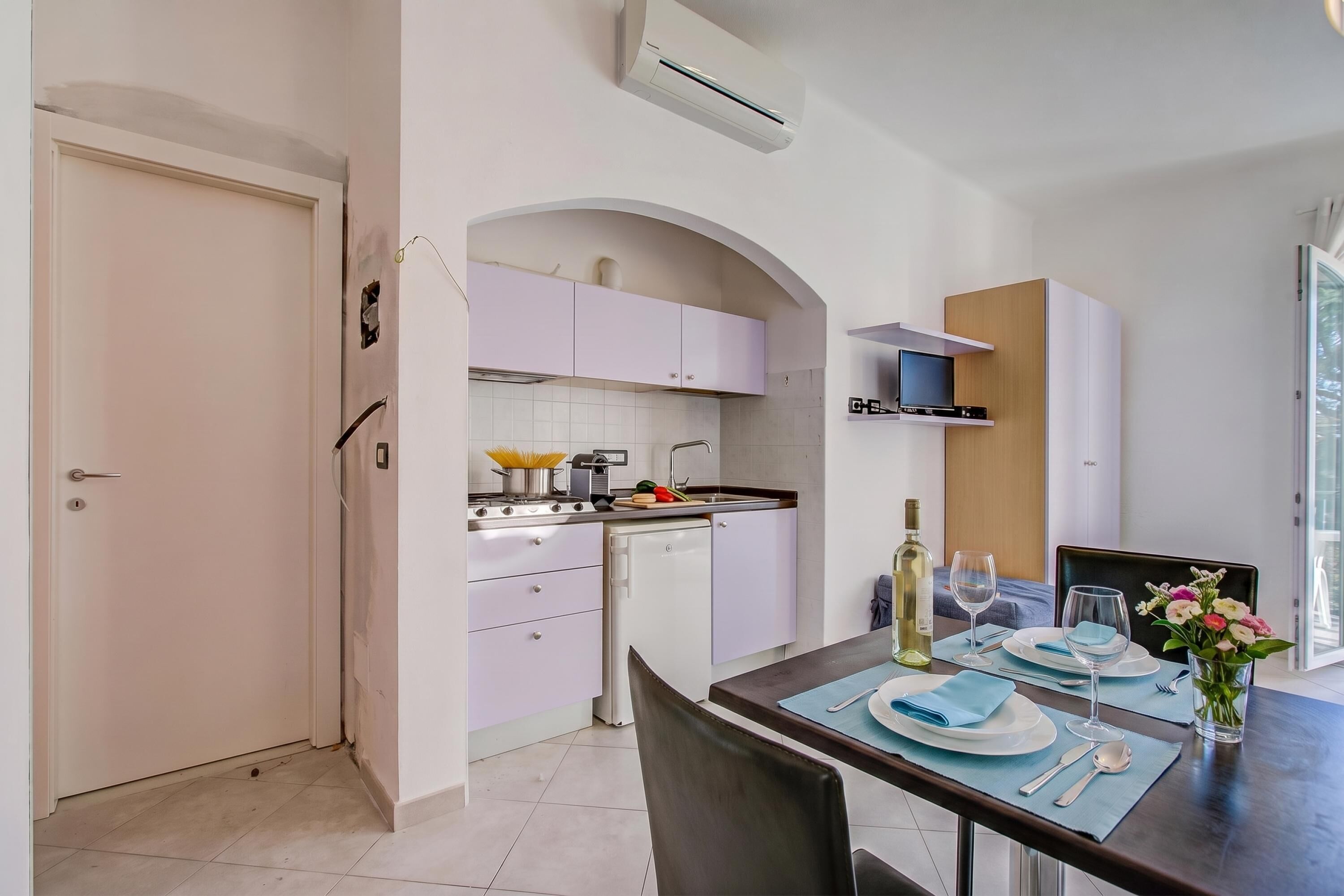 Una cucina moderna con mobili bianchi, forno integrato e zona pranzo per due, con tavolo, sedie e un vaso di fiori.
